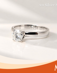 A silver solitaire engagement ring with a round brilliant-cut diamond, displayed on a white surface with a soft shadow. The image includes a '9.